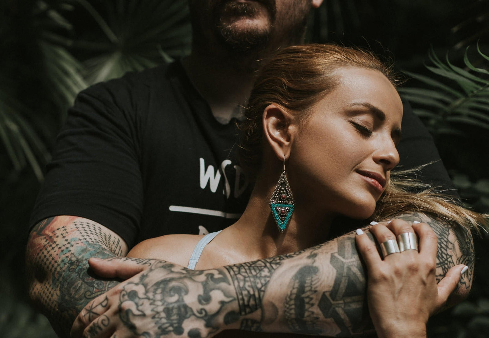 Man with full sleeve black and grey tattoos standing behind a girl with his arms crossed around her chest
