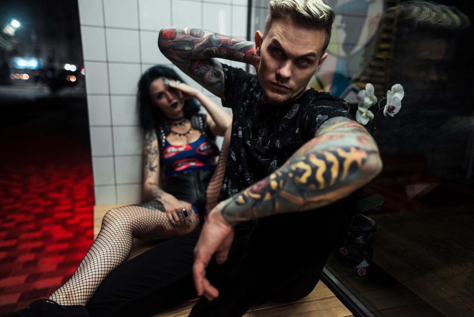 Man with full sleeve color tattoos sitting in front of girl who looks passed out sitting on the floor